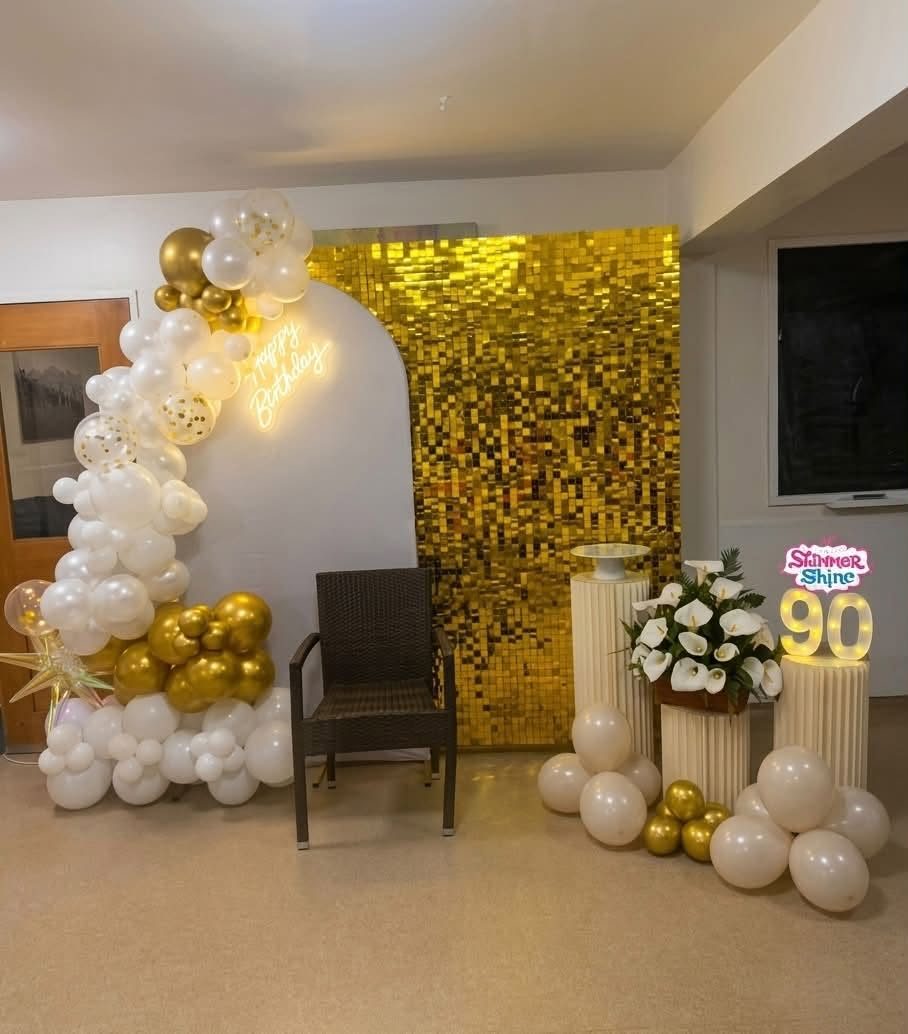 Gold sequin wall with a white arch, balloon garland, neon Happy Birthday sign and flower arrangement set up for a 90th birthday